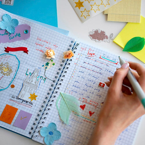 Una persona escribiendo en una libreta de espiral con páginas cuadriculadas, decorada con pegatinas, flores y notas de colores, mostrando una lista de tareas y un título "get for love."