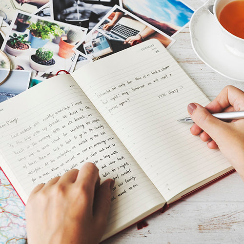 Manos escribiendo en un diario o agenda abierto con páginas rayadas, rodeado de fotografías impresas y una taza de té.
