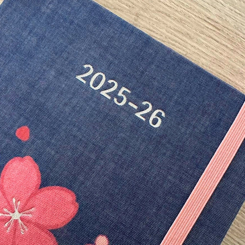 Primer plano de una libreta azul con el grabado "2025-26" y parte de un diseño de flor de cerezo rosa y una banda elástica.