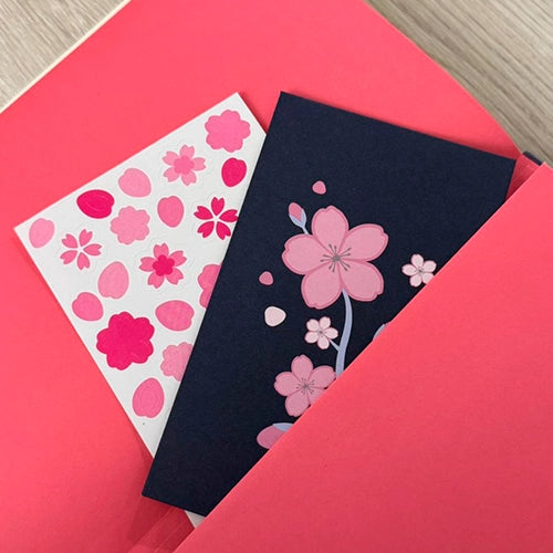 Una tarjeta y una hoja de pegatinas, ambas con motivos de flores de cerezo rosas, sobresaliendo de un bolsillo en una libreta roja.