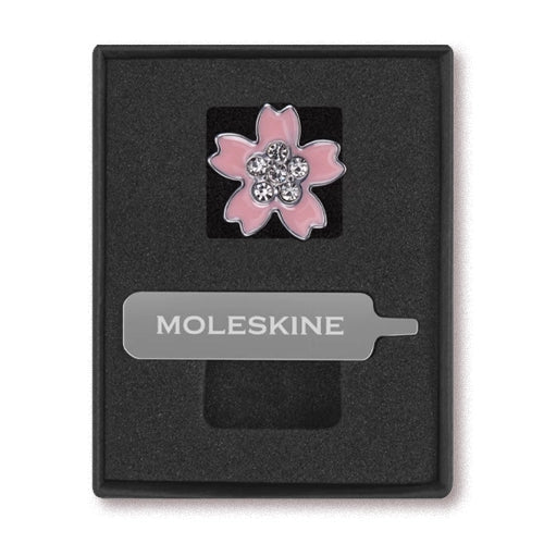 Una caja negra abierta que contiene un pin en forma de flor de cerezo rosa con brillantes y una placa metálica con el logotipo de Moleskine.