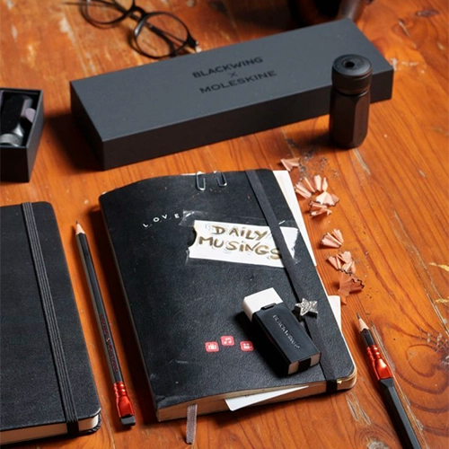 Un cuaderno Moleskine negro abierto, lápices Blackwing, un afilador y gomas de borrar.