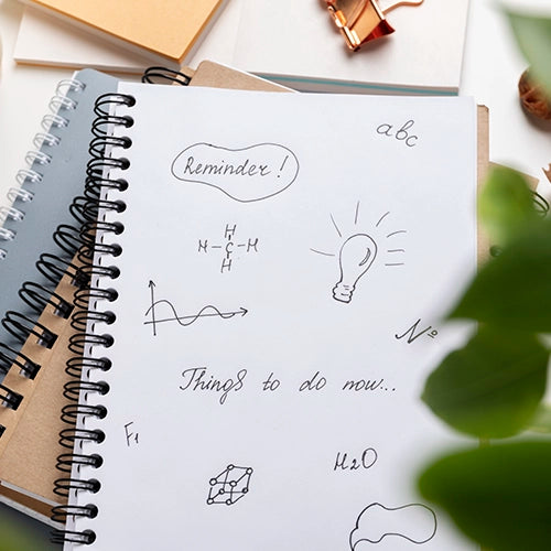 Un primer plano de un cuaderno abierto con dibujos y notas manuscritas, incluyendo un recordatorio, un diagrama de moléculas, una onda, una bombilla y una lista de tareas pendientes.