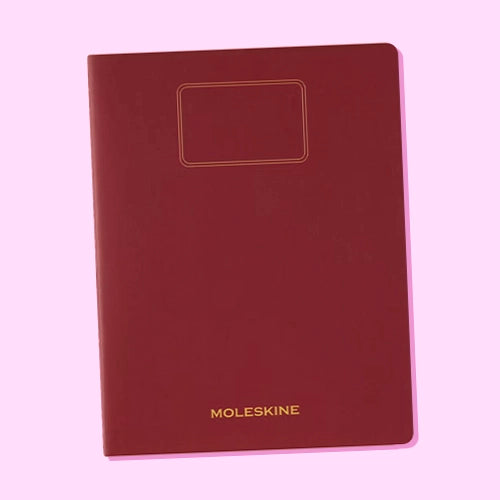 Un cuaderno Moleskine de color borgoña con un recuadro en la portada, sobre un fondo rosa claro.